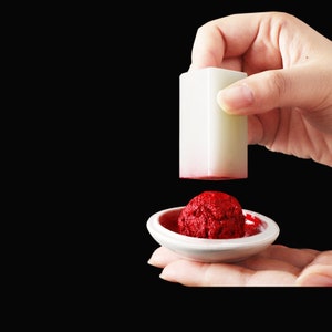 May include: A white rectangular stamp with a red ink pad. The stamp is held above a small, white, round dish filled with red ink. The background is black.