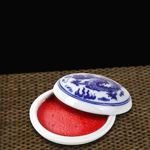 May include: A white ceramic ink pad with a blue dragon design on the lid. The ink pad is open, revealing a bright red ink. The ink pad is on a woven surface.