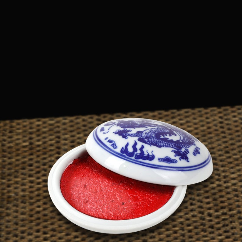 May include: A white ceramic ink pad with a blue dragon design on the lid. The ink pad is open, revealing a bright red ink. The ink pad is on a woven surface.