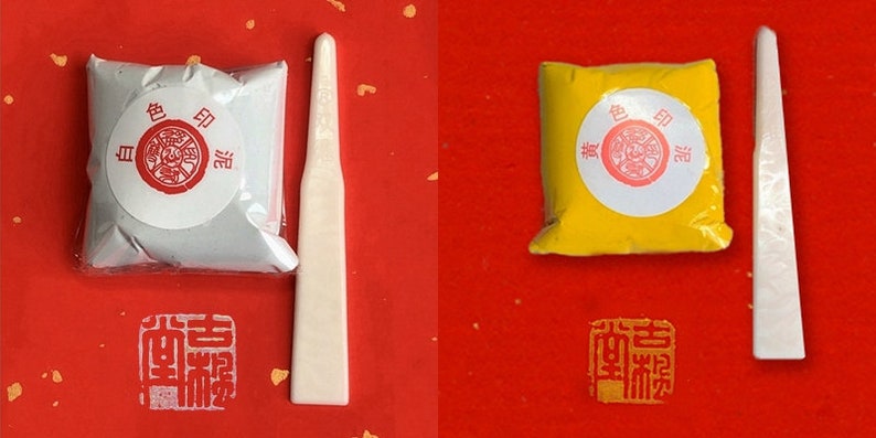 May include: Two small packets of powdered paint, one silver and one yellow, with a white plastic spatula next to each. The packets have Japanese text on them that reads "色 印泥".