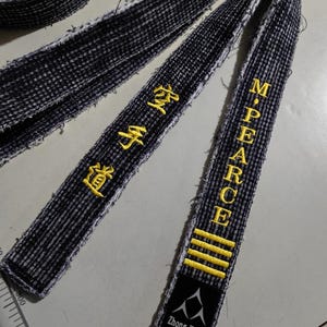 Pu&ograve; includere: Due cinture nere di arti marziali con testo ricamato in giallo. Una cintura ha il testo "Zhong Yuan" e "360cm". L'altra cintura ha il testo "M. PEARCE" e quattro strisce gialle.