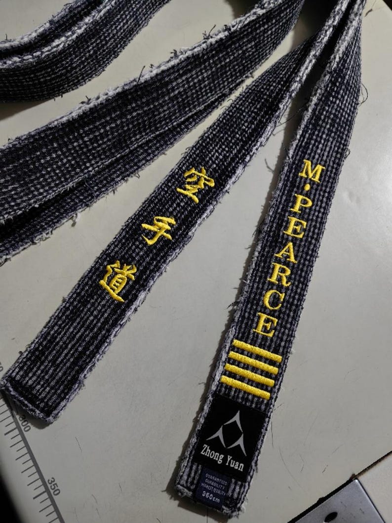 Pu&ograve; includere: Due cinture nere di arti marziali con testo ricamato in giallo. Una cintura ha il testo "Zhong Yuan" e "360cm". L'altra cintura ha il testo "M. PEARCE" e quattro strisce gialle.