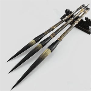 May include: Four calligraphy brushes with black handles and long, tapered bristles. The brushes have a bamboo-like appearance with dark brown and gold accents. They are arranged on a black stand, showcasing their elegant design.