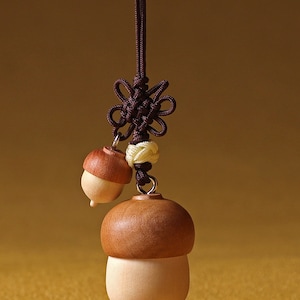 May include: A wooden acorn charm with a brown cord and a knotted tassel. The acorn is made of two pieces of wood, one light brown and one darker brown. The charm is hanging from a brown cord with a knotted tassel.