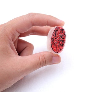 May include: A white oval-shaped stamp with a red and black design. The stamp is held in a hand.
