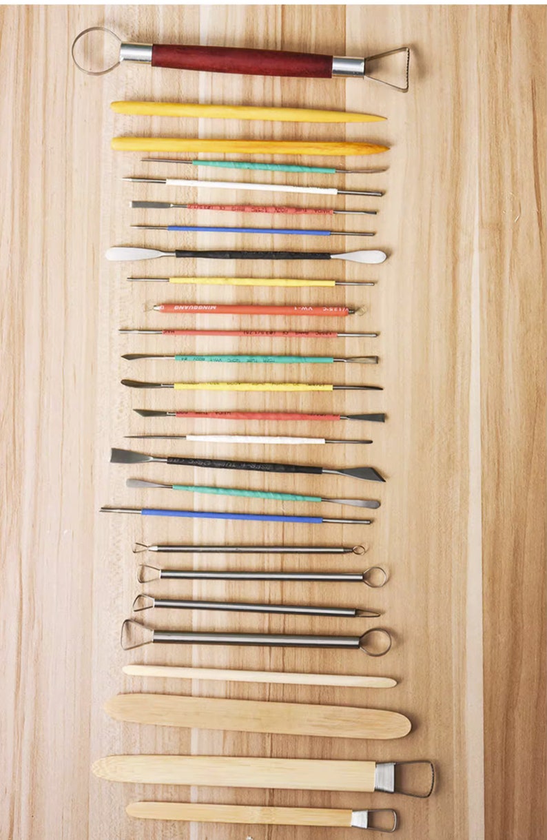 May include: A set of 19 pottery tools, including a loop tool, a wooden paddle, and various sculpting tools with different shapes and sizes. The tools are arranged vertically on a wooden surface.