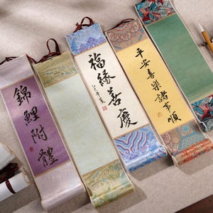 Peut inclure: Une collection de cinq peintures sur rouleau chinoises traditionnelles. Chaque rouleau présente une calligraphie à l'encre noire sur un fond coloré, avec des bordures en tissu décoratives de différentes couleurs et motifs. Les rouleaux sont exposés sur une surface neutre.