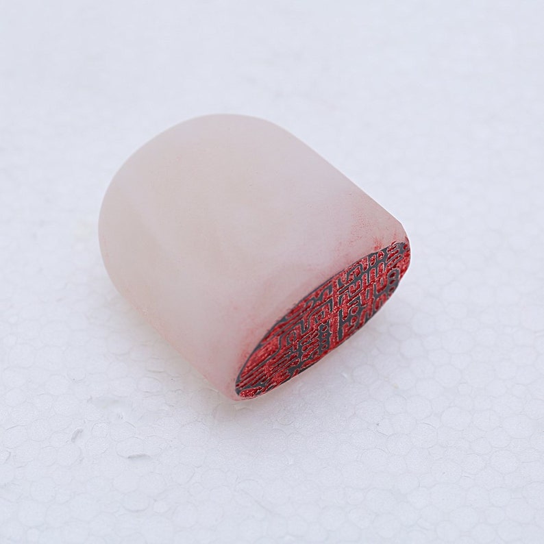 May include: A white, oval-shaped, carved stone seal with a red, detailed design on the bottom.