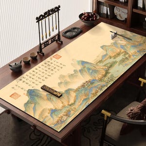 Peut inclure: Un tapis de bureau rectangulaire orné d'une peinture de paysage traditionnelle avec des montagnes bleues et vertes, une rivière et une calligraphie chinoise noire. Le tapis est posé sur un bureau en bois sombre avec un porte-pinceaux et des tasses.