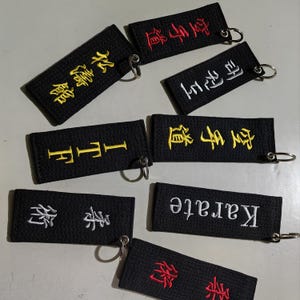 May include: Eight black keychains with different embroidered text. The text includes the words "Karate", "Taekwondo", and Chinese characters. The keychains are on a white surface.
