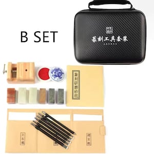 May include: A black carrying case with the text "篆刻工具套装" in white. The case contains a set of tools for carving seals, including a wooden handle with a metal blade, a red ink pad, a small ceramic dish, and a variety of carving tools. The set also includes a book with the text "篆刻研修体系" in black.