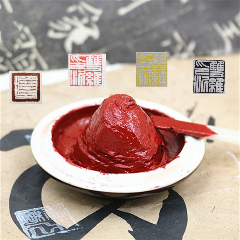 May include: A red ink pad with a small mound of ink in the center. Three square stamps are visible in the background, one red, one yellow, and one black.