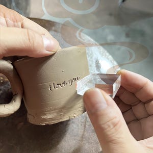 Peut inclure: Une tasse en céramique beige avec une anse, portant l'inscription gravée "I love you". Un outil clair et facetté est tenu près de la tasse, peut-être pour ajouter un motif. La tasse est en cours de fabrication.