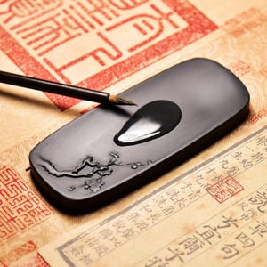 May include: A rectangular black ink stone with a pool of ink and a calligraphy brush. The ink stone features a floral design. The background includes a red stamp and paper with Asian characters.