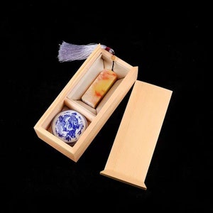 May include: A wooden box with a lid, containing a small, round, blue and white porcelain dish and a rectangular, yellow and white stone. The box has a white tassel attached to the lid.