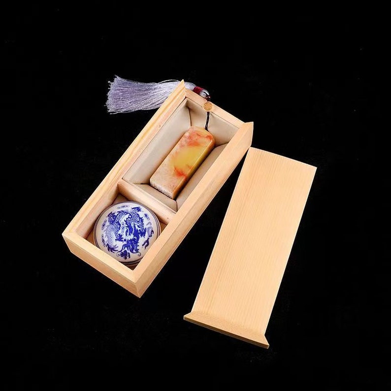 May include: A wooden box with a lid, containing a small, round, blue and white porcelain dish and a rectangular, yellow and white stone. The box has a white tassel attached to the lid.