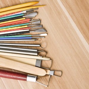 May include: A set of 18 pottery tools, including sculpting tools, a wooden paddle, and a loop tool. The tools are arranged in a fan shape on a wooden surface. The tools are made of metal, wood, and plastic.