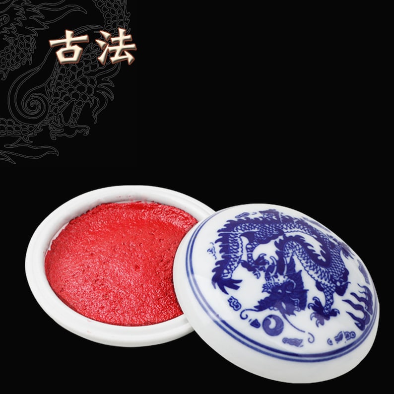May include: A white ceramic ink pad filled with red ink. The lid, decorated with a blue dragon motif, is partially open. The Chinese characters "古法" are visible in the top left corner, set against a black backdrop.