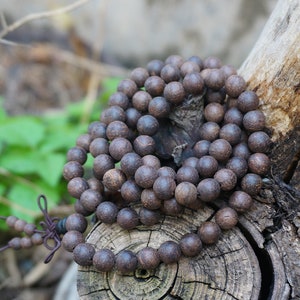 May include: A dark brown wooden beaded necklace with a knotted tassel. The beads are round and smooth, and the necklace is draped over a piece of weathered wood.