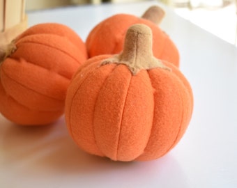 May include: Three orange felt pumpkins with brown stems. The pumpkins are arranged in a row on a white surface.