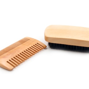 Op de afbeelding: Een houten kam en een houten borstel met zwarte haren. Beide zijn gemaakt van natuurlijk hout en hebben een lichtbruine kleur.