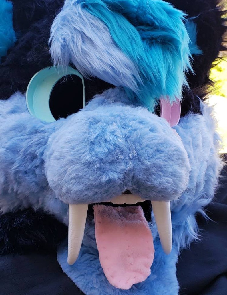 May include: A blue and black furry animal head with a pink tongue sticking out. The animal has large white fangs and a blue and turquoise fur pattern.