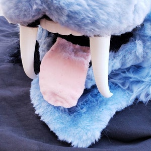 May include: Close-up of a blue plush animal's mouth with large white teeth and a pink tongue sticking out.