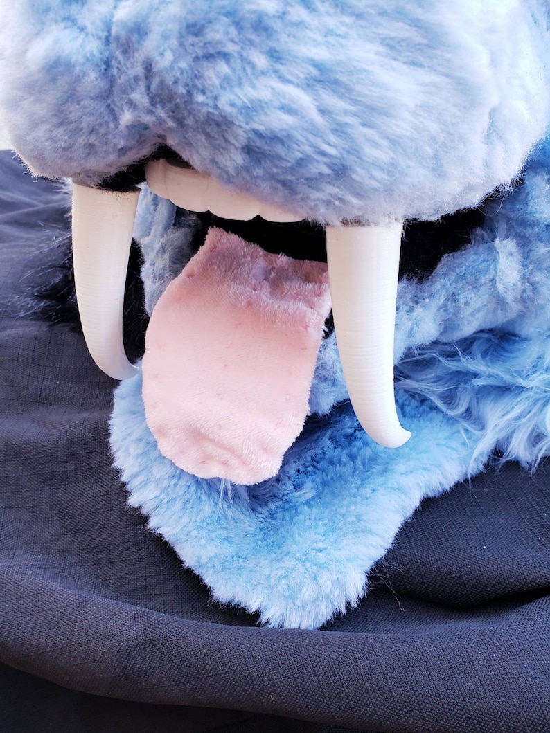 May include: Close-up of a blue plush animal's mouth with large white teeth and a pink tongue sticking out.