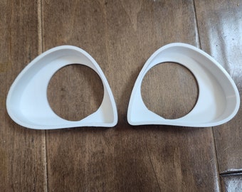 May include: Two white plastic eye-shaped molds with a circular opening in the center. The molds are on a wooden surface.
