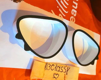 Pode incluir: Um par de óculos de plástico branco com bordas pretas. Os óculos estão sobre uma superfície branca com um pedaço de papel que diz "#classy" com um coração.