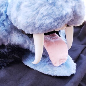 May include: A close-up of a blue plush animal with large white teeth and a pink tongue sticking out. The animal is made of soft, fluffy fur and has a friendly expression.