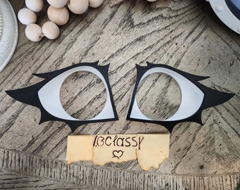 Pode incluir: Dois recortes em forma de olho preto e branco com uma borda dentada. Os recortes estão sobre uma superfície de madeira com uma nota que diz "13C classy" com um símbolo de coração.