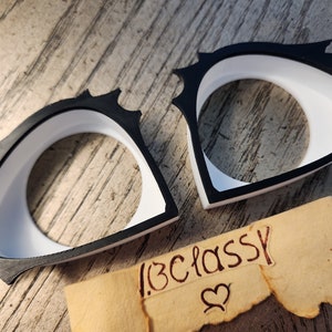 May include: Two black and white plastic eye shapes with a white interior and black exterior. The eye shapes are on a wooden surface with a piece of paper that says "#beclassy" with a heart symbol.