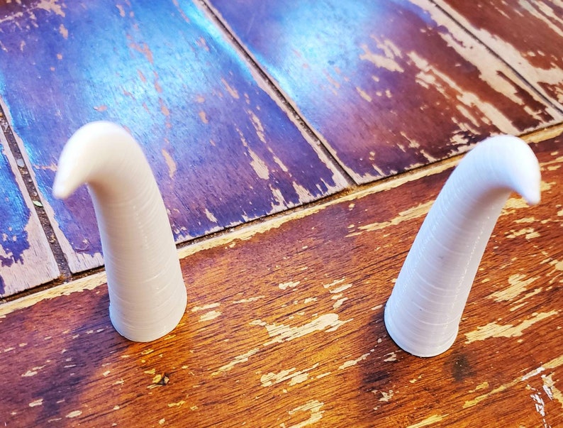 May include: Two white ceramic horns, each with a curved tip, are displayed on a wooden surface.