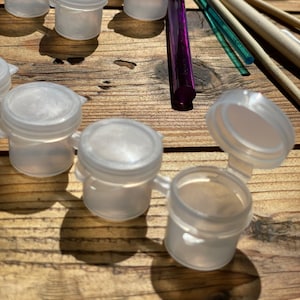 Könnte beinhalten: Nahaufnahme einer Reihe kleiner, transparenter Plastikbehälter mit Deckeln. Die Behälter sind in einem Halbkreis auf einer Holzoberfläche angeordnet. Auf der Oberfläche liegen auch mehrere Pinsel und ein lilafarbener Stift.