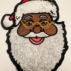 Puede incluir: Parche bordado de un Papá Noel sonriente con barba y bigote blancos, que lleva un gorro rojo y blanco y gafas redondas de montura amarilla. Su piel es marrón.