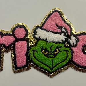 May include: Pink, glitter-edged patch spelling "Grinch". The "Grinch" text has a gold glitter outline. The "i" is replaced with a green Grinch face wearing a pink Santa hat with white trim. The patch is likely for clothing or accessories.