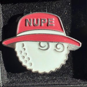 May include: A golf ball marker with a red hat design. The hat has the word "NUPE" in silver letters. The golf ball is white with a silver outline and has a few dimples. The marker is in a black box.