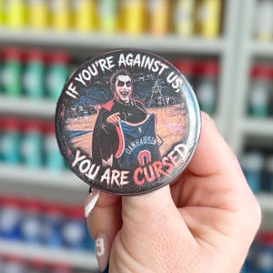May include: A circular pin-back button featuring a graphic of a person with face paint holding a blue jersey with the name DANHAUSEN printed on it. The text IF YOU'RE AGAINST US, YOU ARE CURSED is printed in white and red around the edge.