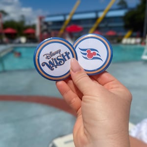May include: Two round pin-back buttons with blue and orange borders. One button features the text Disney WISH in blue script. The other button displays a blue wave graphic with a red Mickey Mouse icon centered above the waves.