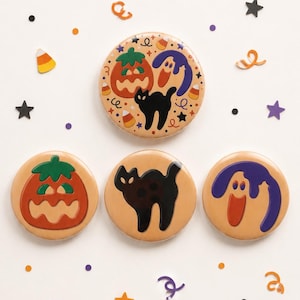 May include: Four round pinback buttons with a tan background. One large button features a pumpkin, a ghost, and a black cat. Three smaller buttons feature a single pumpkin, a black cat, and a ghost respectively, surrounded by confetti.