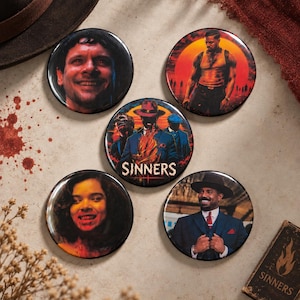 May include: Five circular pin-back buttons featuring imagery from the film Sinners. The central button displays the title SINNERS in white text over a figure in a hat and suit. Other buttons show character portraits, including a smiling person, a person in a tank top, a person with fangs, and a person in a suit and hat.