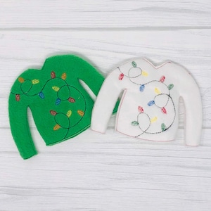 Può includere: Due maglioni di feltro, uno verde e uno bianco, ciascuno ornato di luci natalizie ricamate. Il maglione verde ha un contorno nero, mentre il maglione bianco ha un contorno rosso. Le luci sono di vari colori.