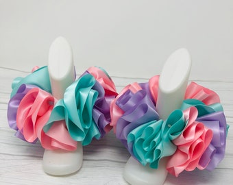 May include: Two pairs of pastel-colored ribbon ankle cuffs. The cuffs are made of pink, purple, and turquoise ribbon and are arranged in a ruffled design. The cuffs are displayed on white mannequin legs.