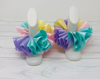 May include: Two white display stands with pastel colored ruffled fabric bows. The bows are made of light blue, lavender, yellow, and pink satin ribbon.