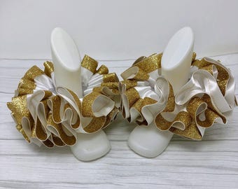 May include: Two white and gold ruffled accessories. Each accessory features a white satin base with gold glitter ribbon accents. The ruffles are layered, creating a voluminous and decorative effect. The accessories are displayed on white forms.