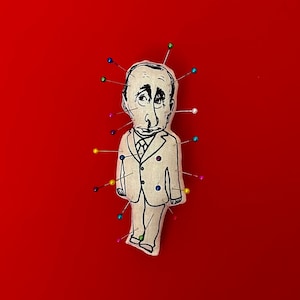 May include: A white fabric doll depicting a man in a suit, with a drawn face, is pinned to a red background with multiple colored pins.