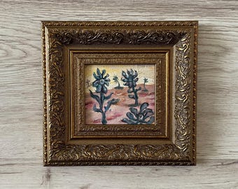 May include: Small framed painting depicting a desert scene with stylized blue flowers. The artwork is set within an ornate, gold-coloured frame. The painting's muted colours and decorative frame give it a vintage feel.