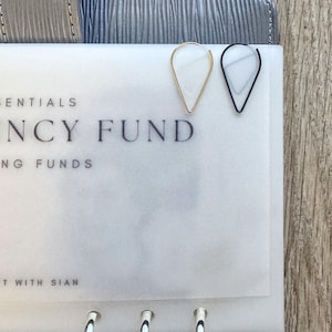 May include: Two teardrop-shaped hoop earrings, one gold and one black, are placed on a white page with the text "ENCY FUND" printed on it. The page is part of a planner with silver rings.