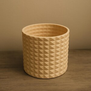Op de afbeelding: Een cilindrische, beige plantenbak met een geometrisch, ruitvormig patroon. De open bovenkant onthult een glad interieur. De plantenbak is gemaakt van een stevig materiaal en staat op een houten oppervlak.
